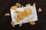 Several ridged potato chips are scattered on a white piece of paper atop a dark wooden surface. The chips are variously whole and broken, giving a casual, laid-back impression.