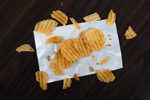 Several ridged potato chips are scattered on a white piece of paper atop a dark wooden surface. The chips are variously whole and broken, giving a casual, laid-back impression.