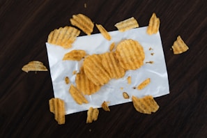 Several ridged potato chips are scattered on a white piece of paper atop a dark wooden surface. The chips are variously whole and broken, giving a casual, laid-back impression.