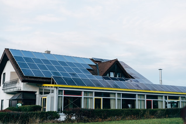 A residential building with a sloping roof covered in solar panels. The structure features multiple windows and is surrounded by well-maintained shrubs. The sky above is cloudy, and a tall, narrow chimney is visible in the background.