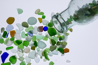 A transparent glass bottle is tipped over, spilling out an assortment of colorful sea glass pieces in various shapes and sizes. The smooth, frosted glass pieces are primarily green, blue, clear, and brown, scattered on a plain white background.