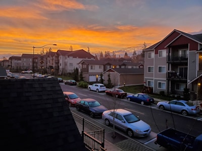 A residential area with multiple apartment buildings and a row of parked cars along the street. The sky is filled with vibrant orange and yellow hues from a sunset. Trees are visible in the background, adding a natural element to the urban setting.