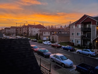 A residential area with multiple apartment buildings and a row of parked cars along the street. The sky is filled with vibrant orange and yellow hues from a sunset. Trees are visible in the background, adding a natural element to the urban setting.