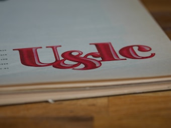 Large, stylized red lettering 'U&lc' on the corner of a page, with the edge of a book in the foreground and a wooden surface underneath.