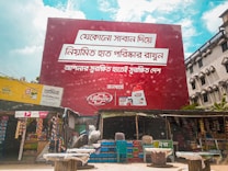 A large red billboard with text in a non-English language dominates the scene, promoting Lifebuoy soap. Below the billboard, there are small shops with various goods displayed, including snacks and household items. The sky is partly cloudy, and there are some buildings visible in the background.