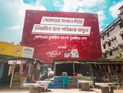A large red billboard with text in a non-English language dominates the scene, promoting Lifebuoy soap. Below the billboard, there are small shops with various goods displayed, including snacks and household items. The sky is partly cloudy, and there are some buildings visible in the background.