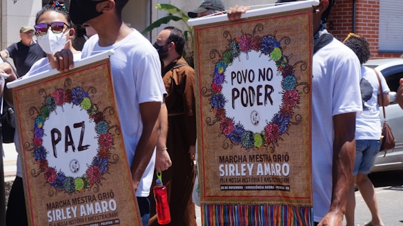 People participate in a public march holding colorful posters with decorative wreaths and text messages advocating for peace and people's power. Participants wear masks, sunglasses, and casual white T-shirts.