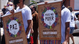 People participate in a public march holding colorful posters with decorative wreaths and text messages advocating for peace and people's power. Participants wear masks, sunglasses, and casual white T-shirts.