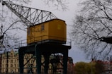 A yellow crane cabin marked with '22 4 TON' is positioned in an industrial or urban area. It is surrounded by bare tree branches, indicating a likely winter or early spring setting. In the background, there are buildings with architectural details suggesting an older style, possibly European.