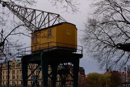 A yellow crane cabin marked with '22 4 TON' is positioned in an industrial or urban area. It is surrounded by bare tree branches, indicating a likely winter or early spring setting. In the background, there are buildings with architectural details suggesting an older style, possibly European.