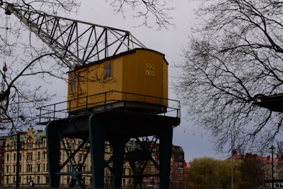 A yellow crane cabin marked with '22 4 TON' is positioned in an industrial or urban area. It is surrounded by bare tree branches, indicating a likely winter or early spring setting. In the background, there are buildings with architectural details suggesting an older style, possibly European.