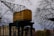 A yellow crane cabin marked with '22 4 TON' is positioned in an industrial or urban area. It is surrounded by bare tree branches, indicating a likely winter or early spring setting. In the background, there are buildings with architectural details suggesting an older style, possibly European.