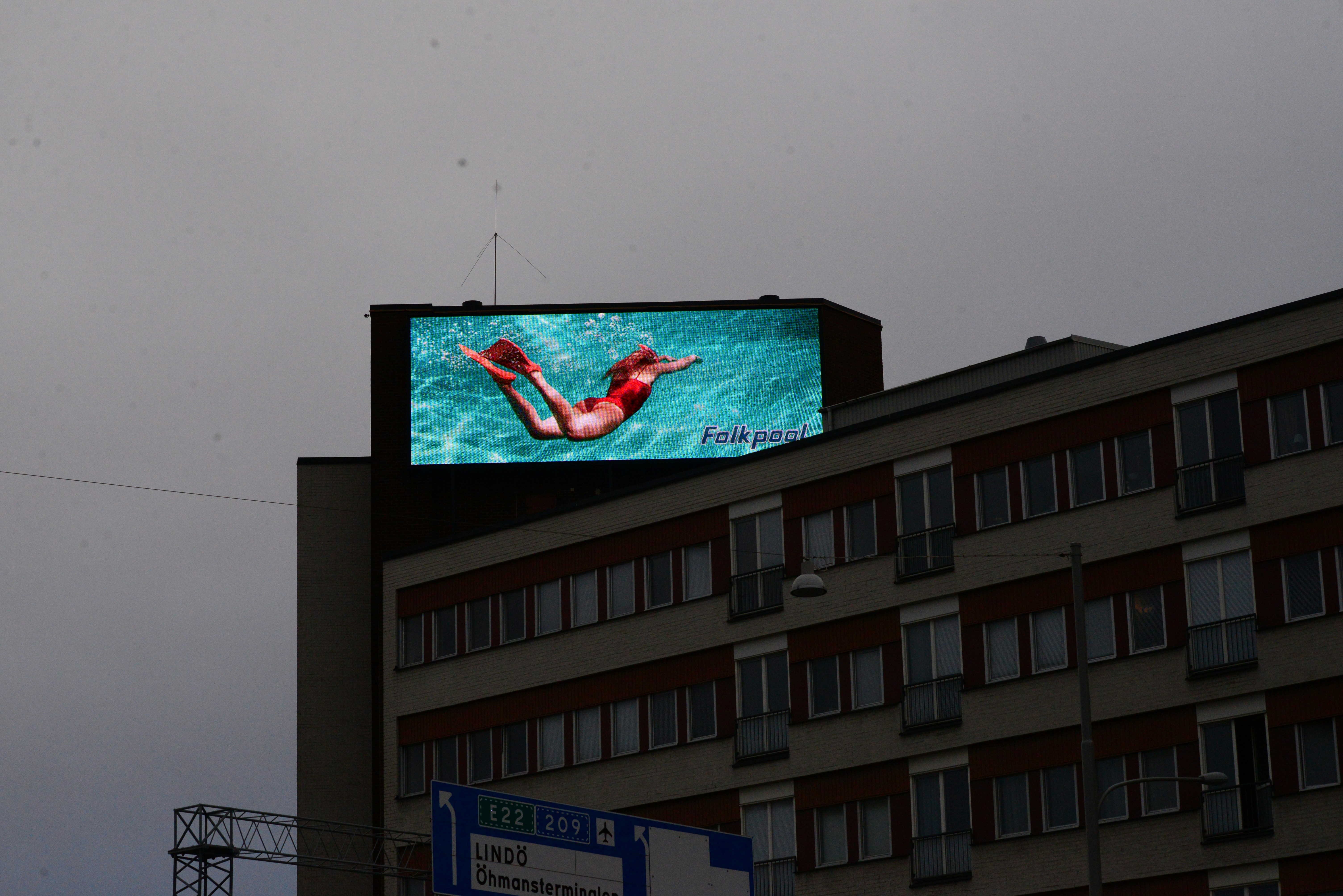 A large digital billboard on top of a multi-story building displays an advertisement featuring a person swimming underwater, wearing bright red swimwear against a vibrant turquoise background. Below, the building features several windows with reddish-brown awnings and some signage near the bottom.