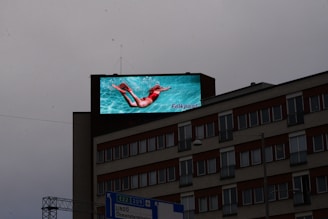 A large digital billboard on top of a multi-story building displays an advertisement featuring a person swimming underwater, wearing bright red swimwear against a vibrant turquoise background. Below, the building features several windows with reddish-brown awnings and some signage near the bottom.