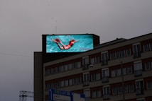 A large digital billboard on top of a multi-story building displays an advertisement featuring a person swimming underwater, wearing bright red swimwear against a vibrant turquoise background. Below, the building features several windows with reddish-brown awnings and some signage near the bottom.