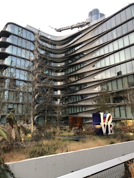 A modern, multi-story building with a distinctive architectural design featuring curved lines and extensive glass windows. The foreground includes a landscaped area with grasses and trees. A large, colorful sculpture of the word 'LOVE' stands prominently on the ground level.