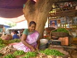 A woman in traditional clothing sits at a market stall under a large tree, sorting through green chilies and ginger. The stall is filled with baskets of fresh vegetables and leafy greens. Behind her are shelves with framed portraits and a vibrant market atmosphere.