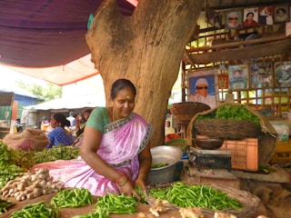 A woman in traditional clothing sits at a market stall under a large tree, sorting through green chilies and ginger. The stall is filled with baskets of fresh vegetables and leafy greens. Behind her are shelves with framed portraits and a vibrant market atmosphere.