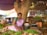 A woman in traditional clothing sits at a market stall under a large tree, sorting through green chilies and ginger. The stall is filled with baskets of fresh vegetables and leafy greens. Behind her are shelves with framed portraits and a vibrant market atmosphere.