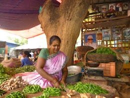 A woman in traditional clothing sits at a market stall under a large tree, sorting through green chilies and ginger. The stall is filled with baskets of fresh vegetables and leafy greens. Behind her are shelves with framed portraits and a vibrant market atmosphere.