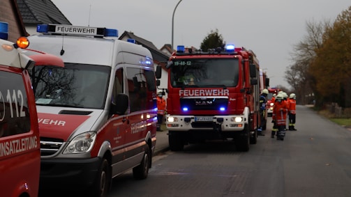 Several fire department vehicles are parked on a residential street, including a fire truck and an emergency response van. A group of firefighters dressed in uniforms and helmets are standing near the vehicles, appearing to be in communication or preparing for action. The scene takes place on an overcast day, with trees without leaves visible in the background.