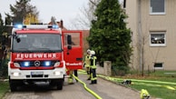 A fire truck with flashing lights is parked on a paved area next to a residential building. Two firefighters in full gear are standing in front of the truck and seem to be engaged in a discussion. Yellow hoses are laid out on the ground leading towards the building, with various equipment scattered around.