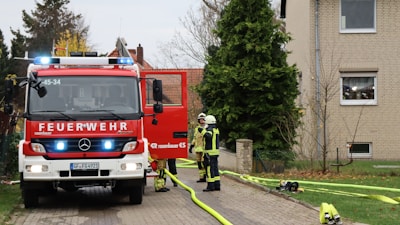 A fire truck with flashing lights is parked on a paved area next to a residential building. Two firefighters in full gear are standing in front of the truck and seem to be engaged in a discussion. Yellow hoses are laid out on the ground leading towards the building, with various equipment scattered around.