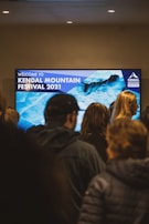 A group of people stand in front of a large digital screen displaying 'Welcome to Kendal Mountain Festival 2021' with a background image of icy blue mountains. The setting appears indoors, with overhead lights illuminating the area.