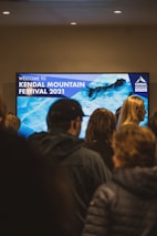 A group of people stand in front of a large digital screen displaying 'Welcome to Kendal Mountain Festival 2021' with a background image of icy blue mountains. The setting appears indoors, with overhead lights illuminating the area.