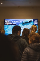 A group of people stand in front of a large digital screen displaying 'Welcome to Kendal Mountain Festival 2021' with a background image of icy blue mountains. The setting appears indoors, with overhead lights illuminating the area.