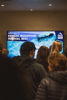 A group of people stand in front of a large digital screen displaying 'Welcome to Kendal Mountain Festival 2021' with a background image of icy blue mountains. The setting appears indoors, with overhead lights illuminating the area.