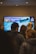 A group of people stand in front of a large digital screen displaying 'Welcome to Kendal Mountain Festival 2021' with a background image of icy blue mountains. The setting appears indoors, with overhead lights illuminating the area.