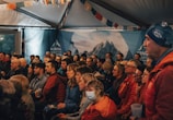 A group of people seated in an indoor setting, attentively watching something off-frame. The room is decorated with colorful banners and a backdrop with mountain imagery. Most individuals are casually dressed and focused, suggesting an engaging event.