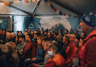A group of people seated in an indoor setting, attentively watching something off-frame. The room is decorated with colorful banners and a backdrop with mountain imagery. Most individuals are casually dressed and focused, suggesting an engaging event.