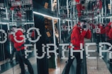 A person wearing a bright red jacket is taking a photograph in a room lined with mirrors, creating multiple reflections. The walls have text that reads 'GO FURTHER' and other smaller text. The scene includes bright lighting from the mirrors and has an industrial feel.