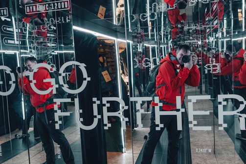 A person wearing a bright red jacket is taking a photograph in a room lined with mirrors, creating multiple reflections. The walls have text that reads 'GO FURTHER' and other smaller text. The scene includes bright lighting from the mirrors and has an industrial feel.