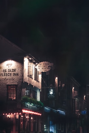 A dimly lit street scene featuring a vintage-style inn with signs advertising 'Ye Olde Fleece Inn' and 'The Fleece Welcomes the Mountain Festival.' The building's façade is illuminated by warm, ambient lighting, and there is a sign with decorative text. Surrounding buildings are partially visible, creating a cozy, inviting atmosphere.