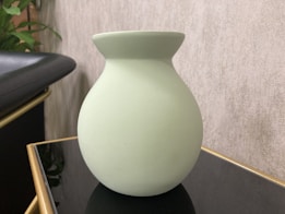 A pale green ceramic vase with a simple, rounded shape sits on a black and gold-edged surface. In the background, there is a textured beige wall and a partial view of green leaves, suggesting plants nearby. The lighting creates a calm and soft atmosphere.
