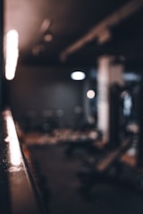 A dimly lit gym setting with blurred exercise equipment in the background and a focus on a reflective surface along the side. The lighting casts a warm glow across the area, enhancing the shadows and creating a moody atmosphere.