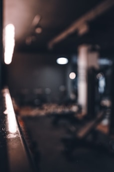 A dimly lit gym setting with blurred exercise equipment in the background and a focus on a reflective surface along the side. The lighting casts a warm glow across the area, enhancing the shadows and creating a moody atmosphere.
