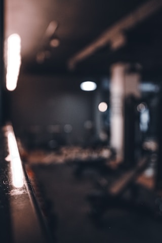 A dimly lit gym setting with blurred exercise equipment in the background and a focus on a reflective surface along the side. The lighting casts a warm glow across the area, enhancing the shadows and creating a moody atmosphere.