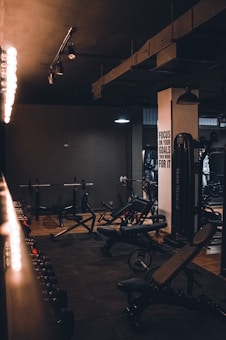 A dimly lit gym with various types of workout equipment including benches, a squat rack, and dumbbells. The walls are dark, and there are industrial-style lights hanging from the ceiling. A motivational sign is visible on one of the walls.