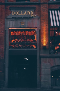 An exterior view of a meat shop with a window display showcasing hanging meat cuts. The sign above the entrance reads 'DOLLARD & Co.', established in 1866. Brick walls and warm lighting create a cozy and inviting atmosphere.