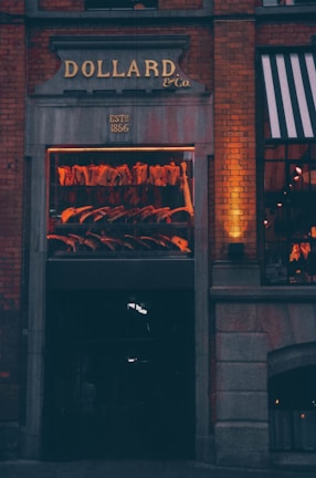 An exterior view of a meat shop with a window display showcasing hanging meat cuts. The sign above the entrance reads 'DOLLARD & Co.', established in 1866. Brick walls and warm lighting create a cozy and inviting atmosphere.