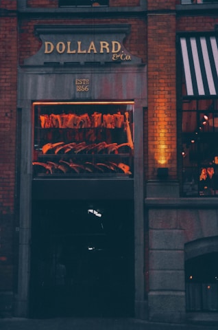 An exterior view of a meat shop with a window display showcasing hanging meat cuts. The sign above the entrance reads 'DOLLARD & Co.', established in 1866. Brick walls and warm lighting create a cozy and inviting atmosphere.