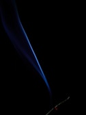 A single, elegant plume of smoke rises, illuminated in shades of blue against a black background. The smoke twists gently upward from a glowing red tip at the bottom, indicating a smoldering object, possibly incense or a cigarette.