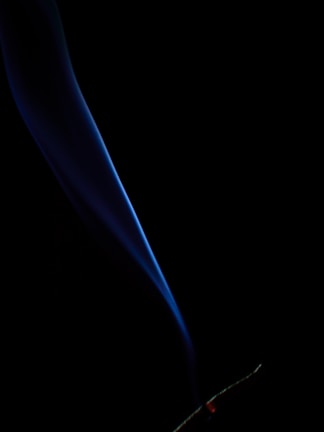 A single, elegant plume of smoke rises, illuminated in shades of blue against a black background. The smoke twists gently upward from a glowing red tip at the bottom, indicating a smoldering object, possibly incense or a cigarette.