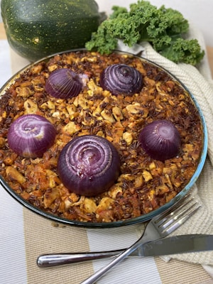 A baked dish in a round glass dish filled with a mixture of rice, vegetables, and four halved red onions. The surface appears crispy and colorful, featuring browned pieces of vegetables and rice. The dish is placed on a tablecloth, and nearby are fresh kale leaves and a whole zucchini, adding a touch of greenery.
