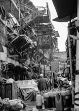 A busy, narrow street filled with various mechanical and industrial items. Numerous car parts and machinery are stacked and hanging on the walls. People are walking through, focused on their tasks, while the surfaces appear wet, suggesting recent rain. The environment is cluttered and lively, indicative of a bustling market or industrial area.