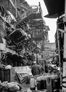 A busy, narrow street filled with various mechanical and industrial items. Numerous car parts and machinery are stacked and hanging on the walls. People are walking through, focused on their tasks, while the surfaces appear wet, suggesting recent rain. The environment is cluttered and lively, indicative of a bustling market or industrial area.